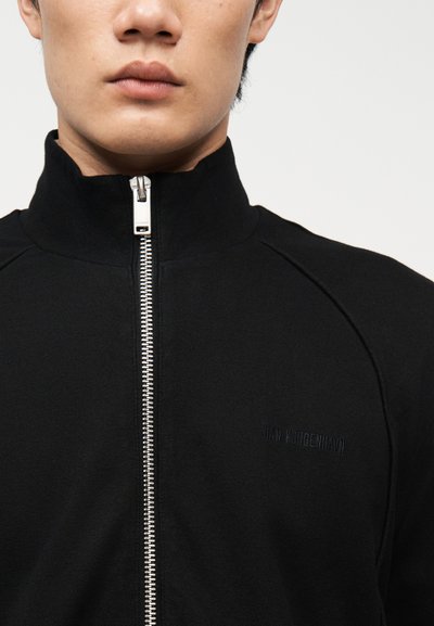 Άνδρας που φοράει μαύρο μπουφάν με φερμουάρ και ψηλό γιακά, όπου φαίνεται το ασημένιο φερμουάρ. Η εικόνα είναι κοντινό πλάνο που δείχνει μόνο το στόμα και τον λαιμό.