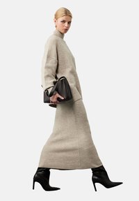 Jasnobeżowy sweter o ciekawym splocie oraz pasująca długa spódnica o prążkowanej fakturze. Modelka ma na sobie czarne szpilki z ostro zakończonym noskiem i trzyma ciemną torebkę.