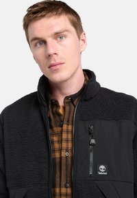 Giacca in fleece nera con colletto alto, tasca sul petto con zip e accenti in materiale liscio a contrasto. Presenta una camicia a quadri sotto.