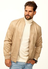 Beige suede bomber jacket with ribbed cuffs and collar, featuring a front zipper and a light-colored sweater underneath. Casual denim jeans.