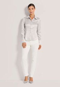 Eine Frau steht vor einem hellen Hintergrund und trägt ein silbernes satinierter Knopfhemd, weiße Hose und graue High Heels.