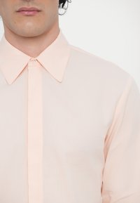 Lys pink langærmet skjorte med klassisk spids krave, knaplukning og glat tekstur. Har diskret syninger langs knappelisten.