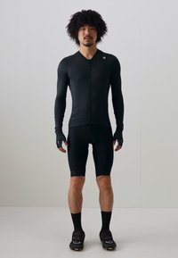 Svart sykkeltrøye med lange ermer og glidelås, kombinert med svarte shorts. Hansker og svarte sykkelsko fullfører den atletiske antrekket.