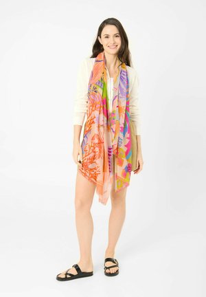 Vrouw draagt een witte trui, kleurrijke bloemen sjaal, beige short en zwarte sandalen, staand en glimlachend tegen een witte achtergrond.