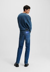 Sweatshirt albastru cu o croială lejeră și guler rotund, asociat cu blugi drepți negri, ținând buzunare la spate și o etichetă de marcă.