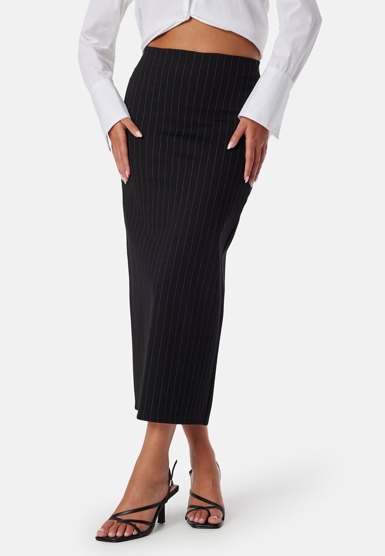 Jupe crayon noire à rayures, longueur mi-mollet, en tissu ajusté. Portée avec une chemise blanche et des escarpins noirs à lanières, mettant en valeur une silhouette élégante.