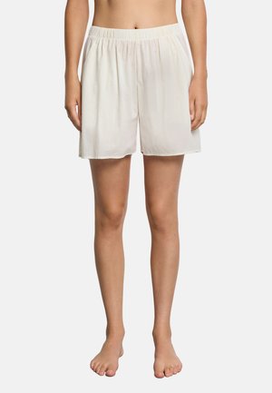 Nainen pukeutunut löysiin, polven mittaisiin valkoisiin shortseihin, joissa on joustava vyötärönauha, seisoo paljain jaloin yksinkertaisen taustan edessä.