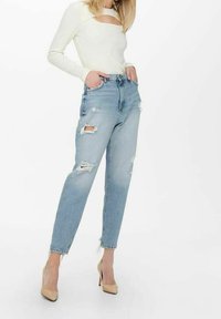 Vaaleansiniset korkeavyötäröiset farkut, joissa on kulutettuja yksityiskohtia, kuten repeämiä ja repsottavat reunat, yhdistettynä valkoiseen pitkähihainen yläosaan ja beigen värisiin korkokenkiin.