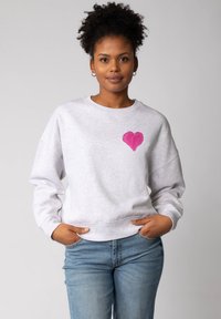 Une jeune femme aux cheveux bouclés porte un sweat-shirt gris clair avec un cœur rose sur la poitrine et un jean bleu, se tenant debout les mains dans les poches.