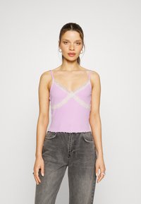 Top de lavanda com rendas cruzadas no peito, apresentando alças finas e uma barra com babados. Usado com calças de ganga cinzentas de cintura alta.