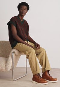 Knit maroon vest with green accents, beige pants, and brown lace-up boots. Sitting on a curved, light-textured chair.