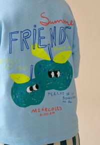 Modrá košile s barevným textem a kreslenými zelenými jablky s očima, obsahuje slova "Summer Friends," "Apples for Sale," a "Miércoles 6:00 AM."