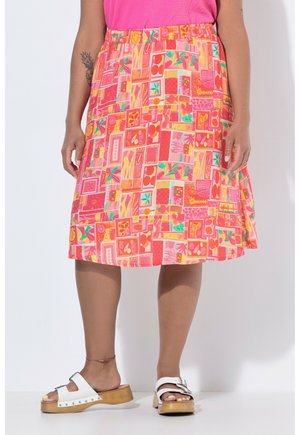 Vrouw draagt een kleurrijke rok met geometrisch patroon, roze top, witte houten platformsandalen en een enkelbandje, staand tegen een witte achtergrond.