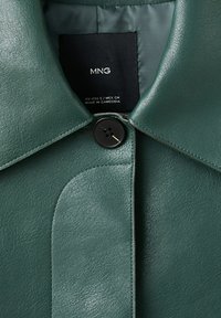 Gulerul unei jachete din piele verde închis, cu un nasture negru înnodat și o etichetă interioară neagră ce afișează detalii despre marcă și mărime.