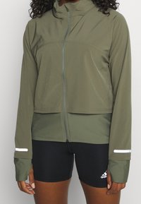 Olivgrön idrottjacka med framzip, hög krage och reflexdetaljer. Har lång ärmar med tumhål och en kortad design.