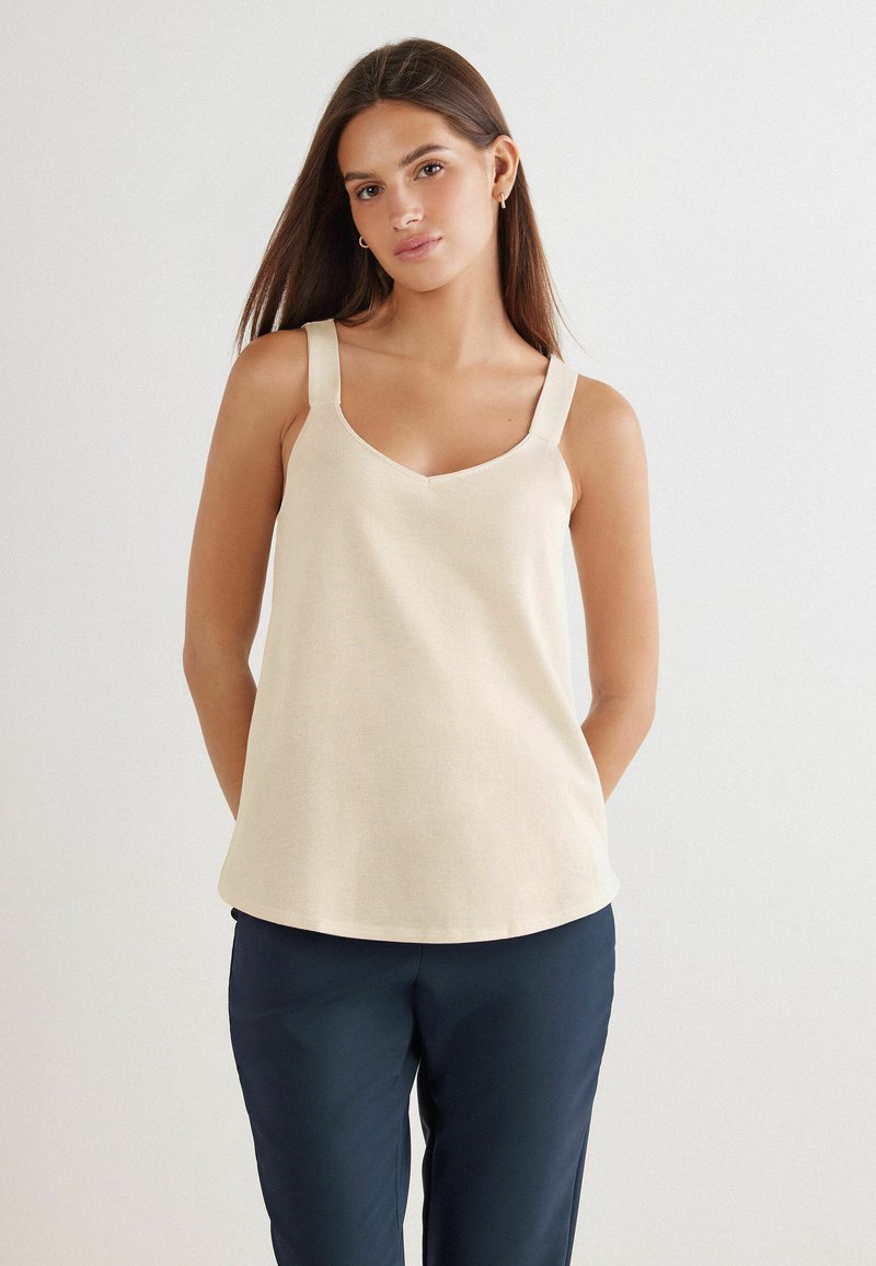 Beiges ärmelloses Oberteil mit V-Ausschnitt, breiten Trägern und weicher Textur, kombiniert mit dunkelblauen Hosen. Einfaches, klares Design ohne Muster.