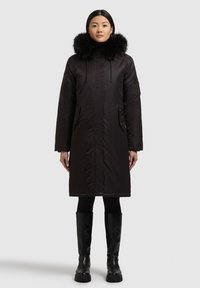 Musta parkatakki, jossa on turkisvuorattu huppu, vetoketjullinen etuosa, sivutaskut ja keskipitkä malli. Yhdistetty mustiin istuviin housuihin ja polven korkeisiin saappaisiin.