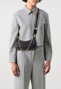 Bolso de hombro de cuero negro con un acento de cadena dorada y correa ajustable, que presenta un logo audaz. La persona lleva una camisa de cuadros y pantalones.