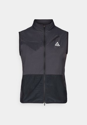 Svart ermeløs vest med glidelås, høy krage, to brystlommer med glidelås, og en liten trekantet logo på venstre bryst.