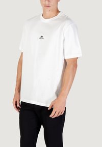 Camiseta blanca de manga corta con cuello redondo, que presenta un pequeño logo negro en el pecho. La tela parece suave y ligera.