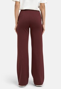 Frau in hochgeschnittenen, weiten burgunderroten Hosen und einem weißen Kurzarmshirt, steht mit entspannten Händen an den Seiten.