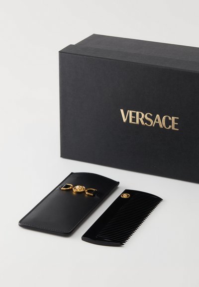 Svart kam med gullaksenter, hviler ved siden av et svart lær-etui med et gullemblem, plassert ved siden av en svart boks merket "VERSACE."