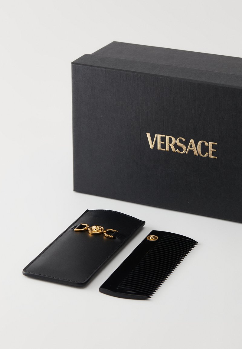 Schwarzer Kamm mit goldenen Akzenten, neben einer schwarzen Lederetui mit goldem Emblem liegend, neben einer schwarzen Schachtel mit der Aufschrift "VERSACE" platziert.