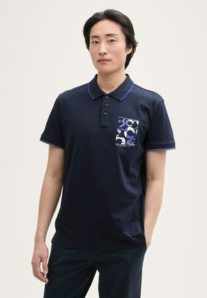 Marineblauwe polo gemaakt van katoen met korte mouwen, een drie-knopenknopsluiting en een patroonzak op de linkerborst met blauwe en witte ontwerpen.