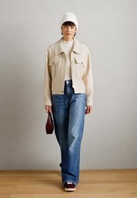 Chaqueta beige corta con dos bolsillos frontales, usada sobre una blusa blanca y jeans de pierna ancha azules. Combinación de zapatillas burdeos y un bolso rojo pequeño.