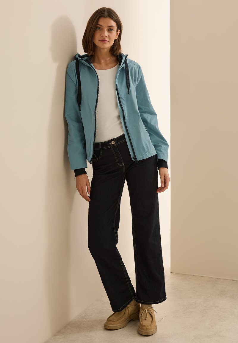 Veste à capuche bleu clair avec des accents noirs, t-shirt blanc, jeans foncés avec des coutures contrastées, et chaussures beiges. Tissu lisse, coupe décontractée.