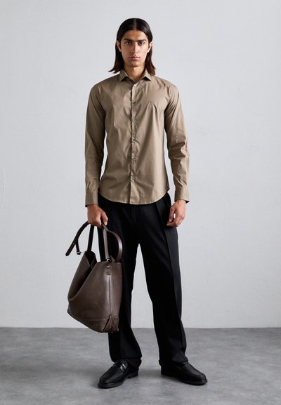 Chemise marron à manches longues avec des boutons, pantalon sombre et chaussures noires texturées. Tient un sac fourre-tout en cuir marron foncé. Couleur unie, design subtil.