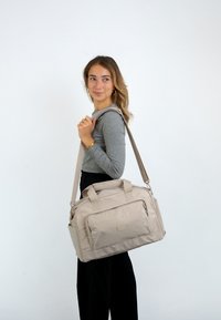Béžová textilná kabelka typu duffel s dvojitými hornými ušami a odnímateľným ramenným popruhom. Má prednú zipsovú kapsu a hladký povrch.
