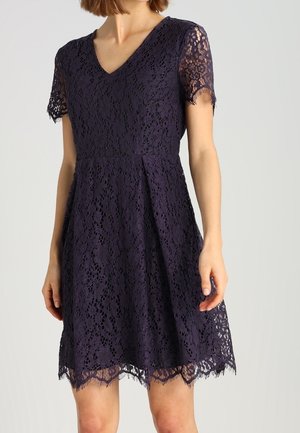 Kobieta ubrana w ciemnofioletową koronkową sukienkę o długości do kolan, z krótkimi rękawami, dekoltem w serek i falowanym dołem, stojąca na jednolitym tle.