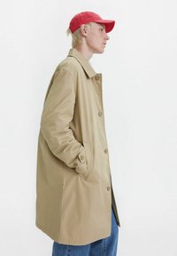 Cappotto beige di lunghezza ginocchio con chiusura a bottoni e tasche laterali; tessuto liscio; indossato sopra jeans blu; abbinato a un cappellino rosso.