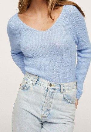 Nainen pukeutuneena vaaleansiniseen V-kaula-aukkoseen neulottuun villapaitaan ja vaaleanpesuun olevaan korkeavyötäröisiin farkkuihin, kädet lepäävät selän takana.