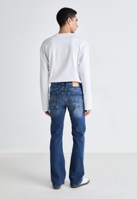 Camisa blanca de manga larga, jeans de denim azul con un ligero desgaste y costuras distintivas en los bolsillos traseros. Calzado informal completa el look.