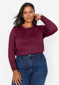 Haut à manches longues bordeaux avec décolleté festonné et accents en dentelle complexes sur les manches, associé à un jean en denim bleu taille haute.