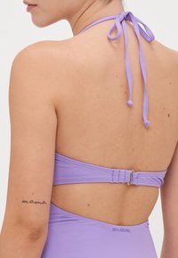 Maillot de bain violet à dos nu, avec une fermeture à crochet en argent et des bretelles à nouer. Texture lisse avec un branding minimaliste à la taille.