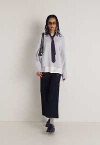 Ανοιχτό γκρι υπερμεγέθη πουλόβερ σε στρώσεις πάνω από ένα λευκό πουκάμισο, συνδυασμένο με σκούρες φαρδιές παντελόνες και μαύρες loafers. Αξεσουάρ περιλαμβάνουν γυαλιά ηλίου και μια μικρή τσάντα.