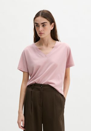 Młoda kobieta z długimi ciemnymi włosami, ubrana w jasnoróżowy T-shirt z dekoltem w serek, włożony do ciemnobrązowych spodni z wysokim stanem, na białym tle.
