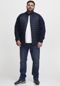 Jūrinės spalvos kviltuota striukė su aukštu apykakle ir ilgomis rankovėmis, derinama su mėlynomis džinsomis ir tamsiais sportbačiais. Minkšta medžiaga su paminkštintu paviršiumi.