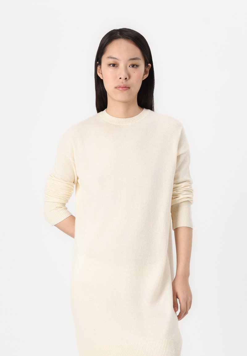 Kremowa sukienka z długim rękawem o luźnym kroju i prążkowanymi mankietami, wykonana z miękkiego materiału, z okrągłym dekoltem i prostym wzorem.