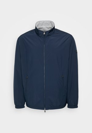 Tamsiai mėlyna lengva striukė su aukštu apykakle, priekyje užtrauktuku ir elastingomis rankogaliais. Turi šonines kišenes ir lygią, blizgią tekstūrą.