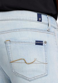 Jasnoniebieskie dżinsy z tylną kieszenią, zdobioną falistym złotym ściegiem oraz ciemnoniebieską metką z białym napisem.