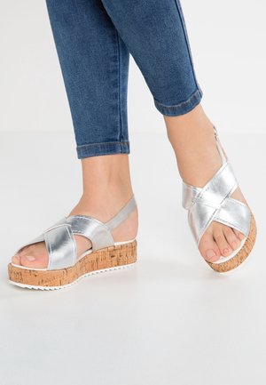 Sølvmetallic kilehæle sandaler med korsremme, kork platform og hvid gummisål. Åben bagside design, minimalistisk stil.