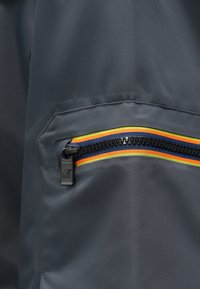 Sivi tkaninski jopič z žepom na zadrgo, okrašen s trikobarvno črto rumene, oranžne in modre barve. Gladek otip, sodoben dizajn.