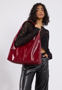 Sac à main en cuir verni rouge au design décontracté et grandes ouvertures, tenu par une personne portant un top en tricot texturé noir et un pantalon en cuir.