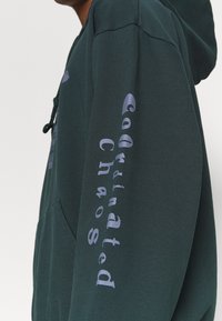 Tmavě zelená mikina s kapucí se stahovací šňůrkou a boční kapsou. Na pravém rukávu je nápis „Coordinated Chaos�“ světle fialovou barvou. Měkká textura.