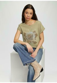 T-shirt vert olive clair avec un imprimé graphique, associé à un jean bleu et des bottines beige. Le t-shirt a des manches courtes et une coupe décontractée.
