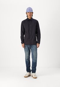 Camisa de manga larga a rayas negras, jeans de mezclilla oscuros y zapatillas blancas. Lleva una gorra de color púrpura claro, de pie contra un fondo liso.